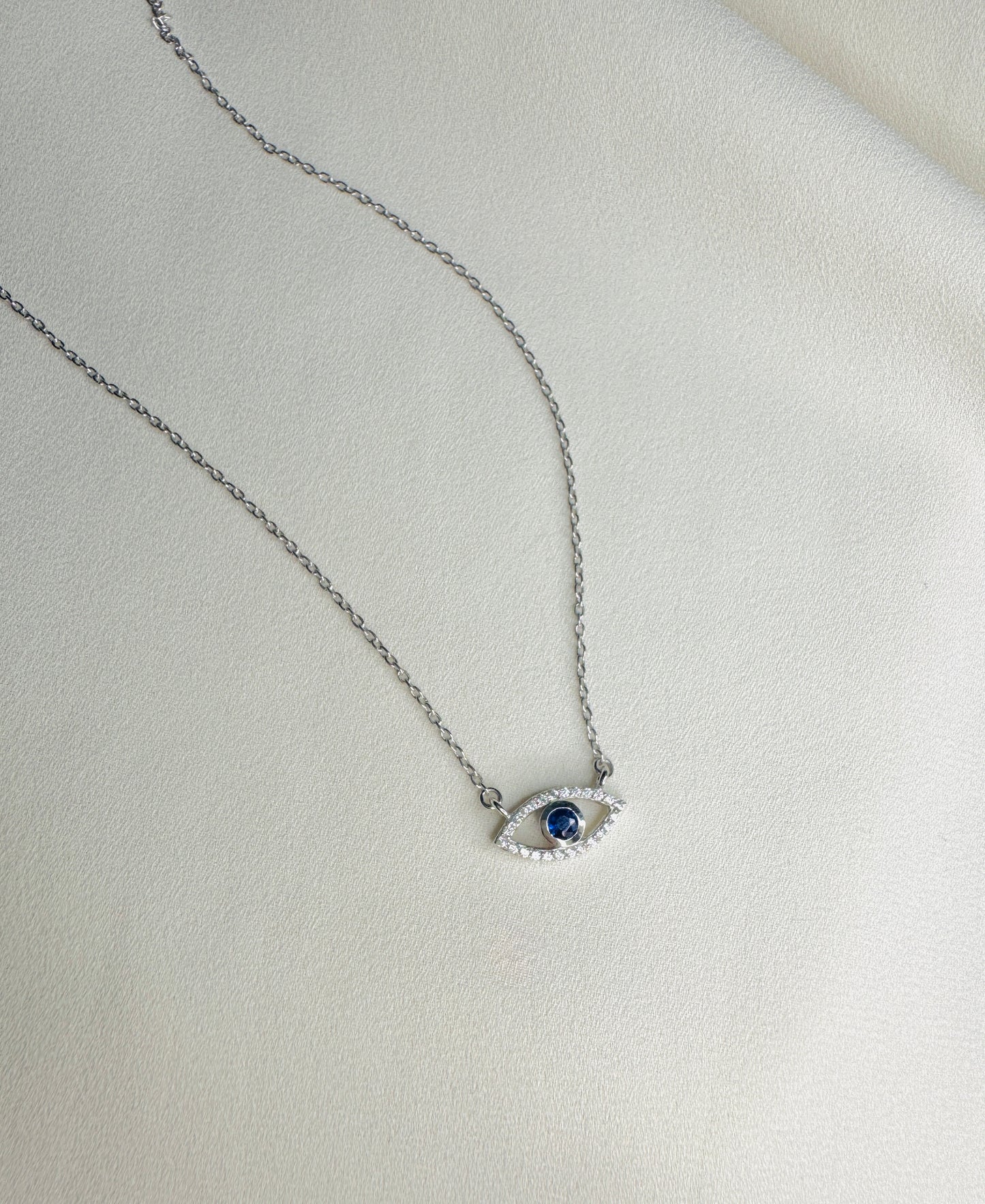 A silver-toned chain necklace with an evil eye pendant featuring a dark blue center stone and small clear stones around it, displayed on a textured off-white surface.