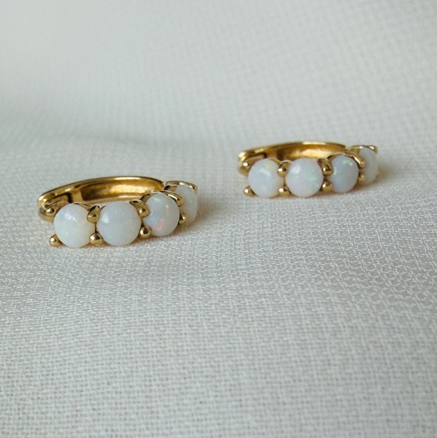 A pair of gold huggie hoop earrings, each featuring three round white opal-like stones, displayed on a textured off-white fabric background.