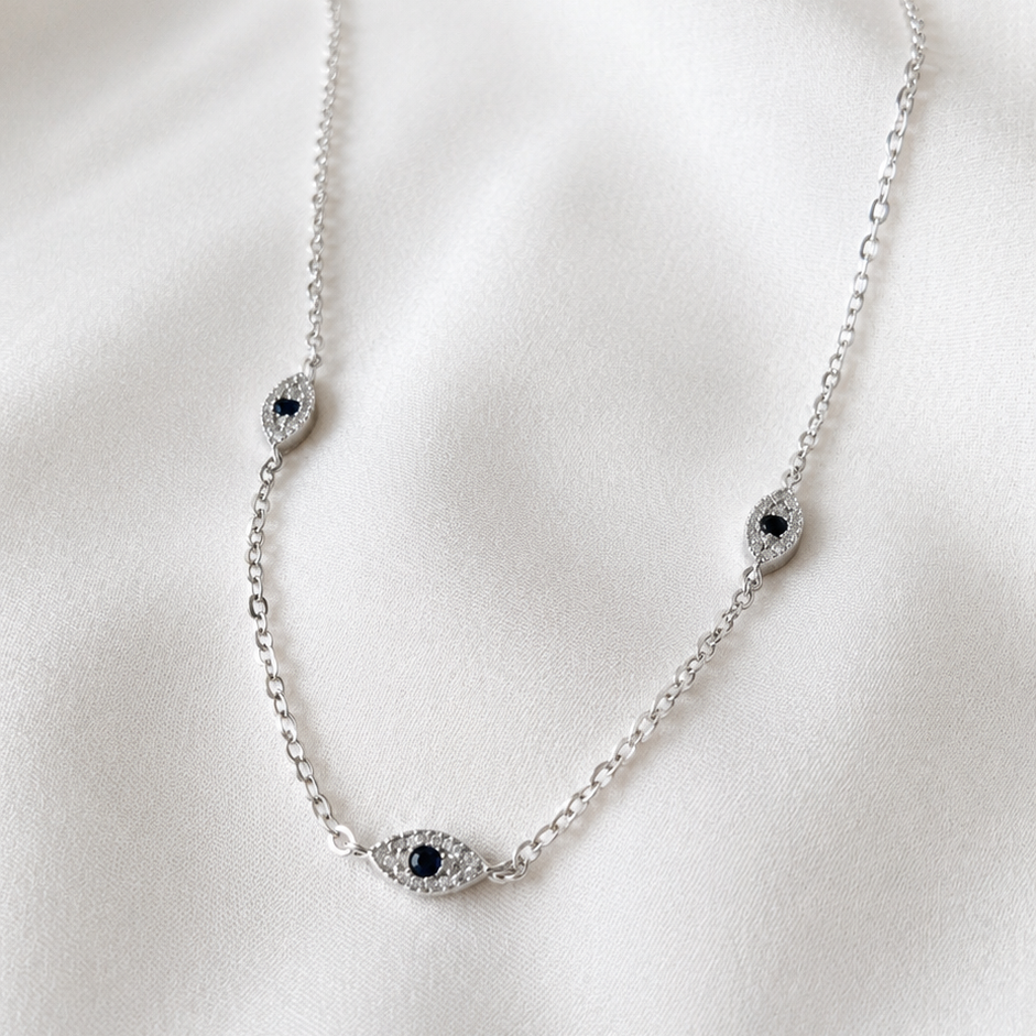A delicate silver chain necklace featuring three evil eye charms with small black centers, displayed on a soft, draped white fabric background.