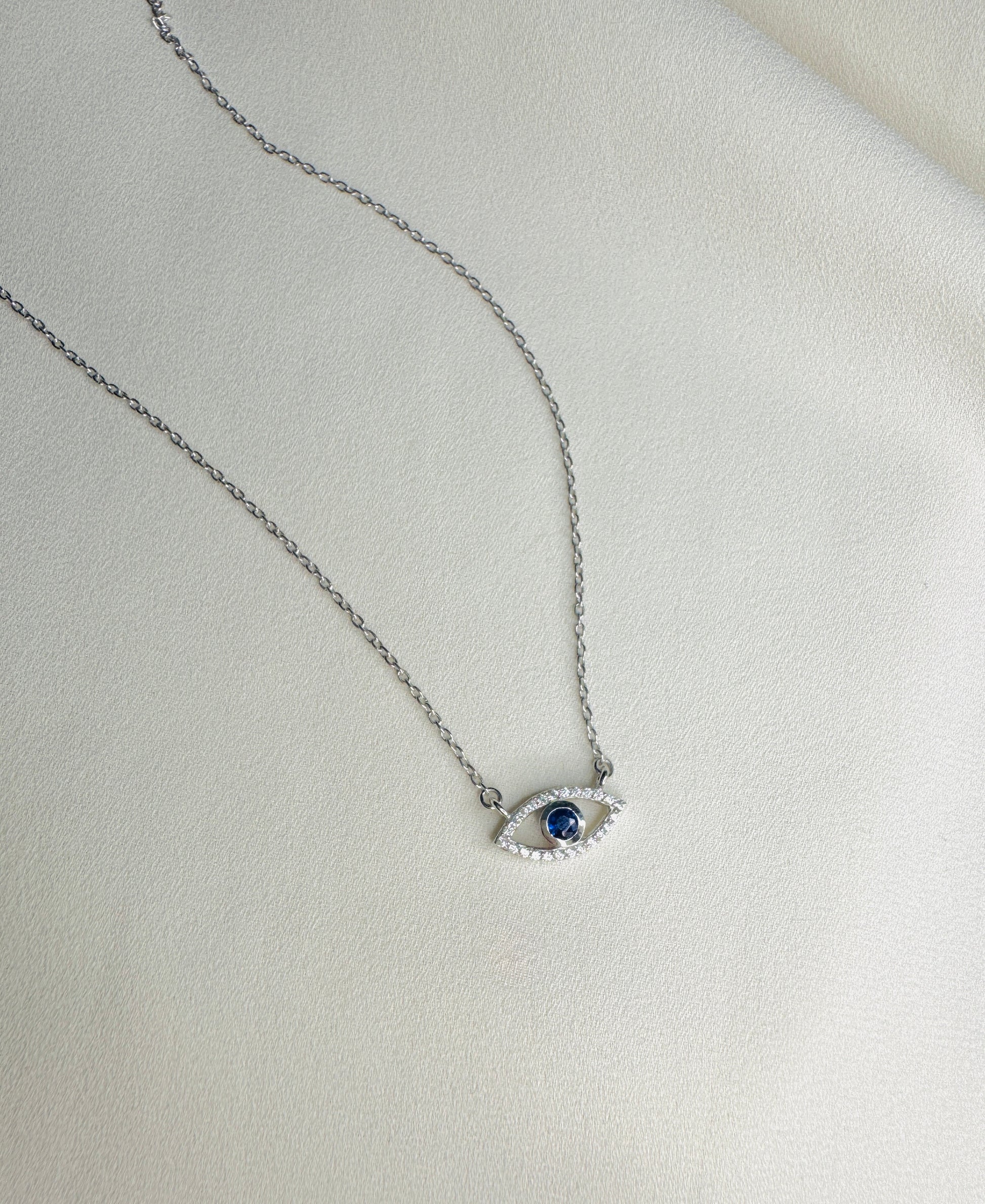 A silver-toned chain necklace with an evil eye pendant featuring a dark blue center stone and small clear stones around it, displayed on a textured off-white surface.