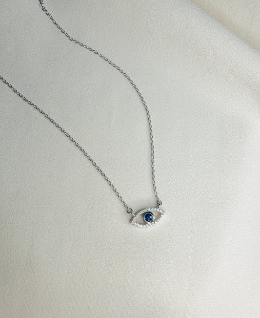 A silver-toned chain necklace with an evil eye pendant featuring a dark blue center stone and small clear stones around it, displayed on a textured off-white surface.