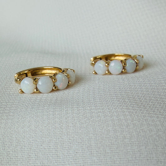 A pair of gold huggie hoop earrings, each featuring three round white opal-like stones, displayed on a textured off-white fabric background.